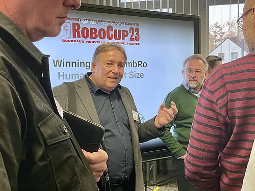 Eine Person steht vor einem gro&szlig;en Bildschirm und spricht zu anderen Menschen. Auf dem Bildschirm steht RoboCup23.