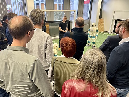 Eine Person steht nehmen einem dem Menschen &auml;hnlichen Roboter und spricht zu einer Gruppe von Menschen.